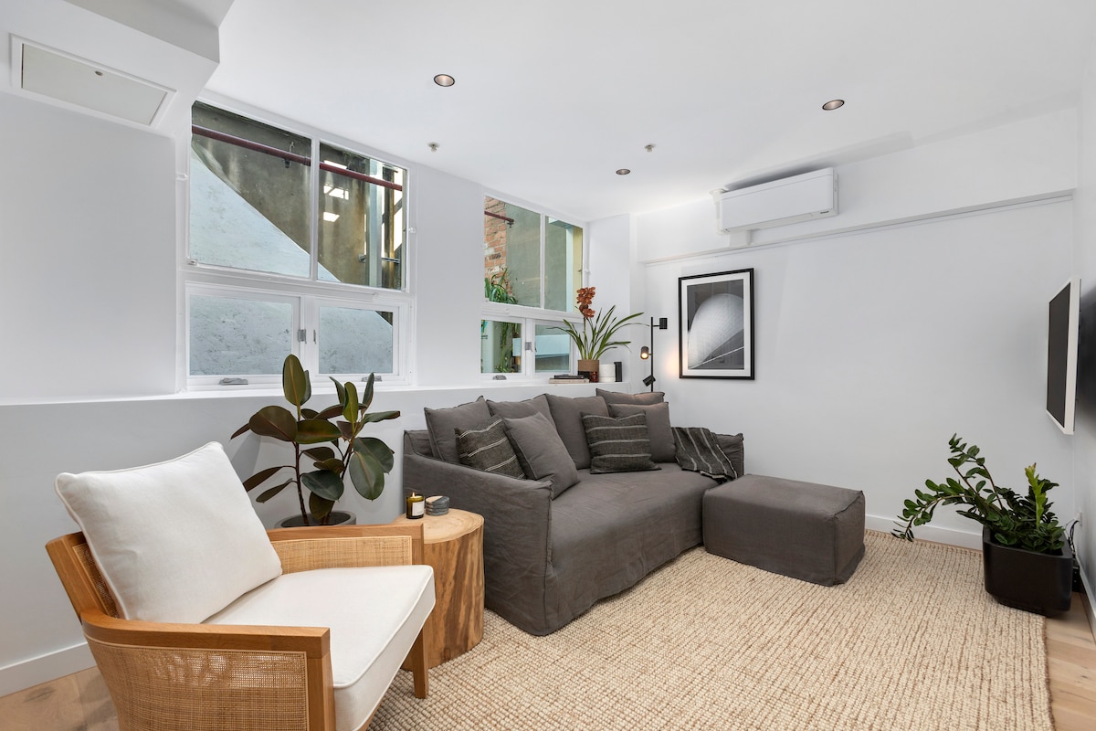 A comfortable seating area is created with a spacious dark gray sofa adorned with neutral cushions. A light wooden armchair and a small side table are positioned nearby. Lush green plants are placed throughout, and large windows allow natural light to fill the room.