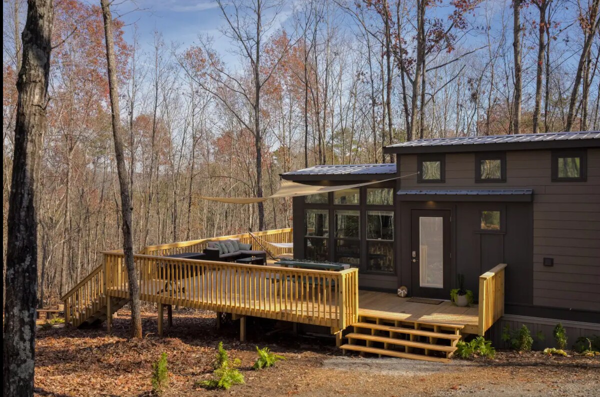 A tiny home is nestled among trees, featuring a spacious wooden deck with a hot tub. Large windows offer views of the wooded surroundings. A gentle slope leads to the entrance, enhancing accessibility. The deck is partially shaded by a canopy, inviting relaxation in nature.