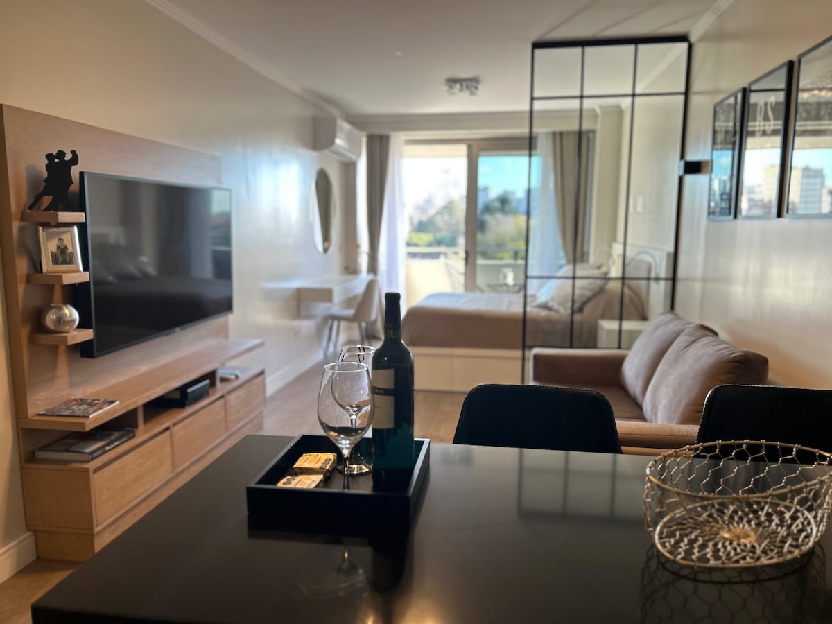 A modern studio layout is visible, featuring a cozy seating area with a brown sofa and a TV unit. A glass table is set with a wine bottle and glass. The background reveals a spacious bed and large windows allowing natural light to fill the room.