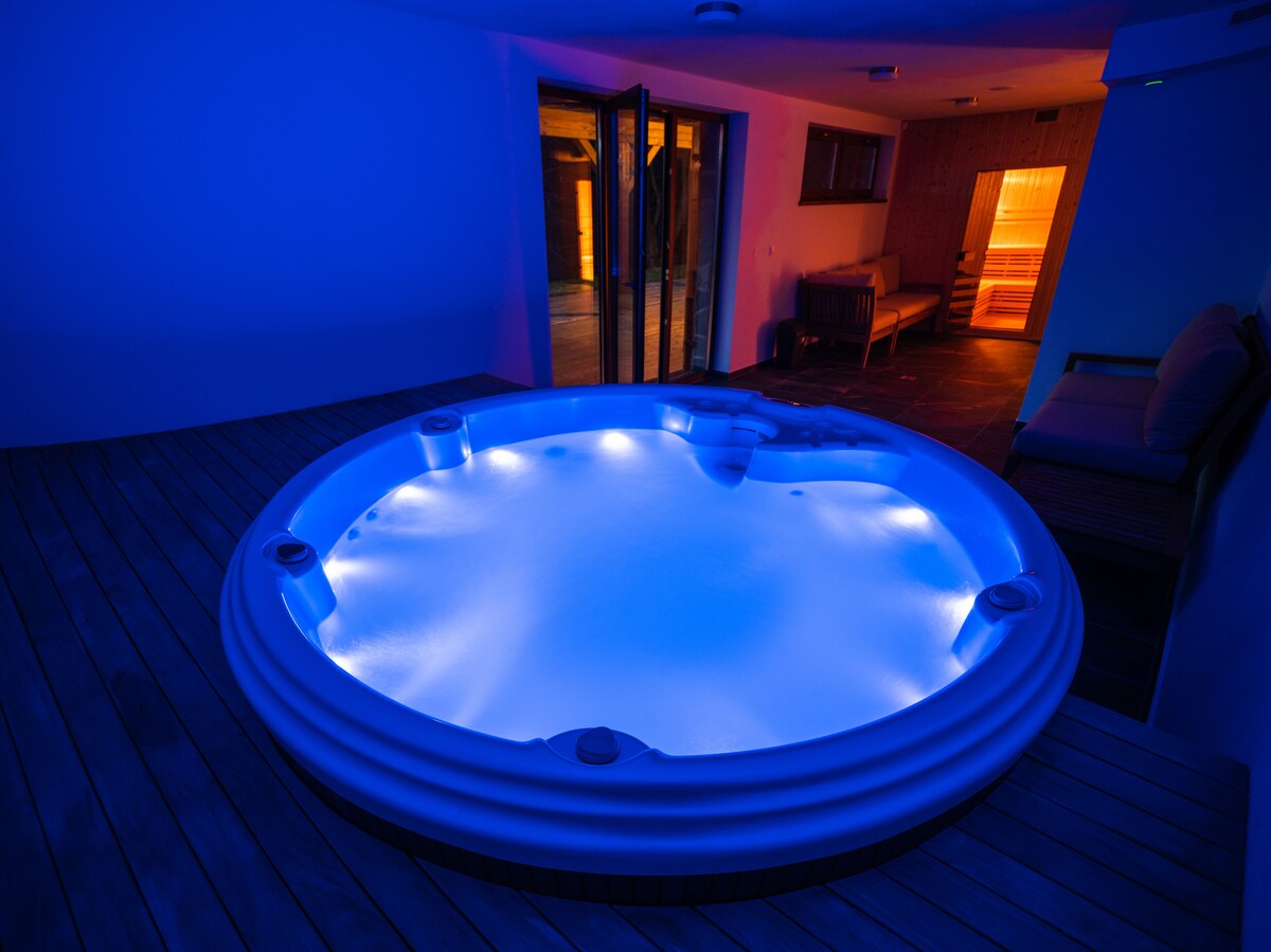 A spacious jacuzzi features softly glowing blue lights, set against a backdrop of wooden decking. The room includes a glimpse of a sauna and lounge area, creating a serene wellness retreat.
