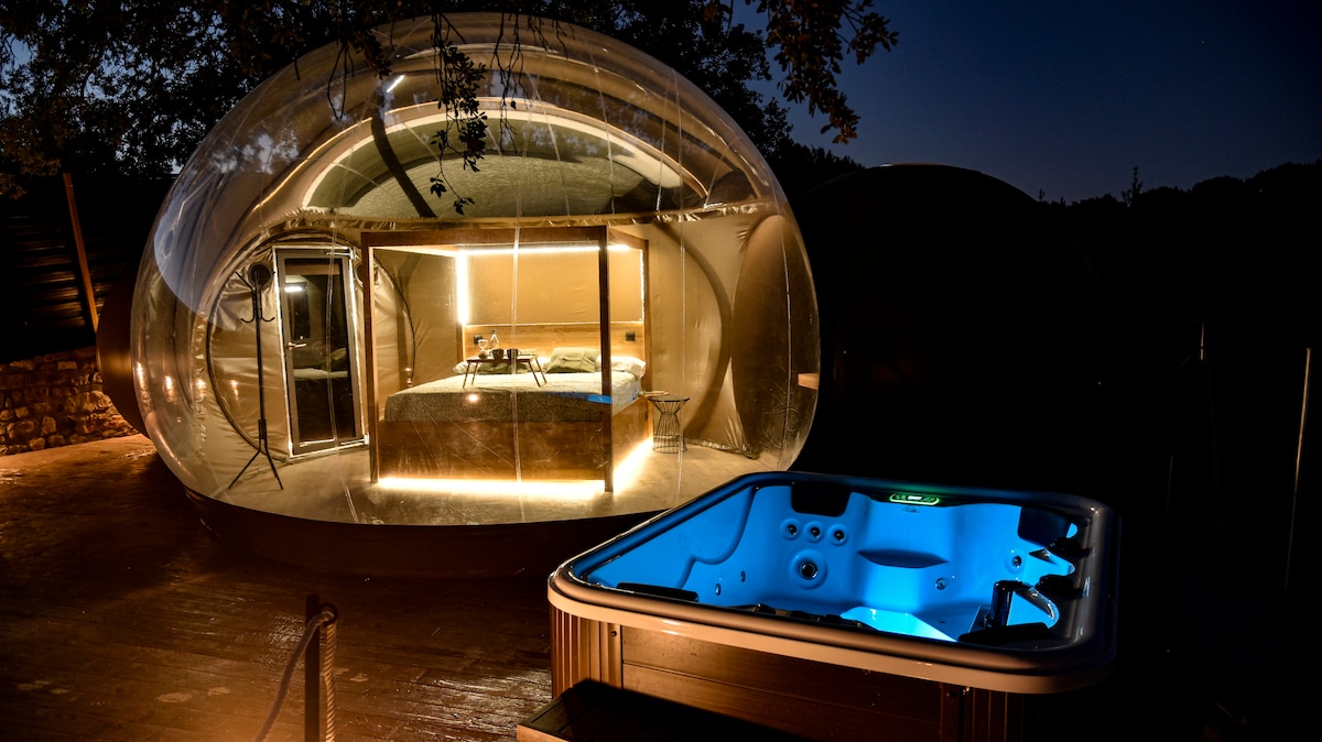 A transparent bubble structure is set against a nighttime backdrop, featuring a cozy sleeping area illuminated with soft lighting. A hot tub with blue underwater lights is positioned on the wooden deck, enhancing the unique outdoor experience.
