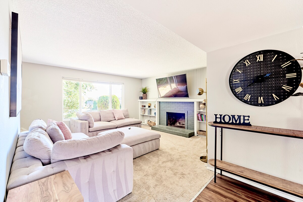 A cozy living room is featured, containing a light-colored sectional sofa and a coffee table. A large television is mounted above a decorative fireplace. Natural light fills the room through a spacious window, with decorative shelves and a wall clock adding character.