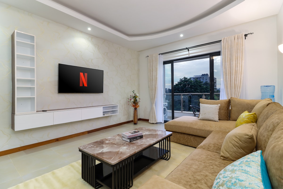 The spacious living area features a large, plush sectional sofa arranged around a sleek marble coffee table. A wall-mounted TV displays the Netflix logo, while large windows allow natural light to fill the room, offering views of the outside balcony.