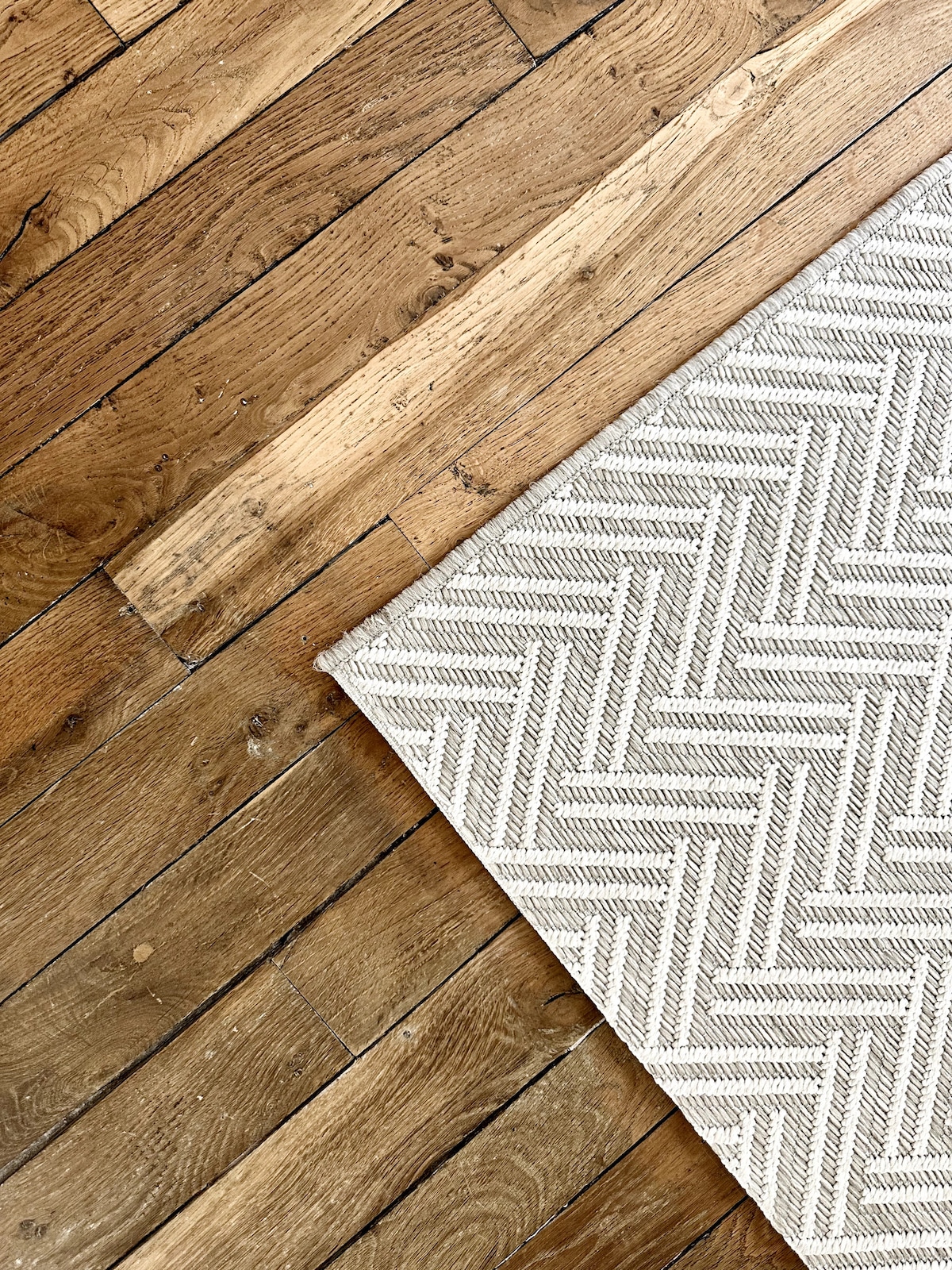 A section of wooden flooring features a blend of rich tones, showcasing natural grain patterns. A textured area rug with a subtle herringbone design lies partially across the floor, providing a soft contrast to the polished wood.