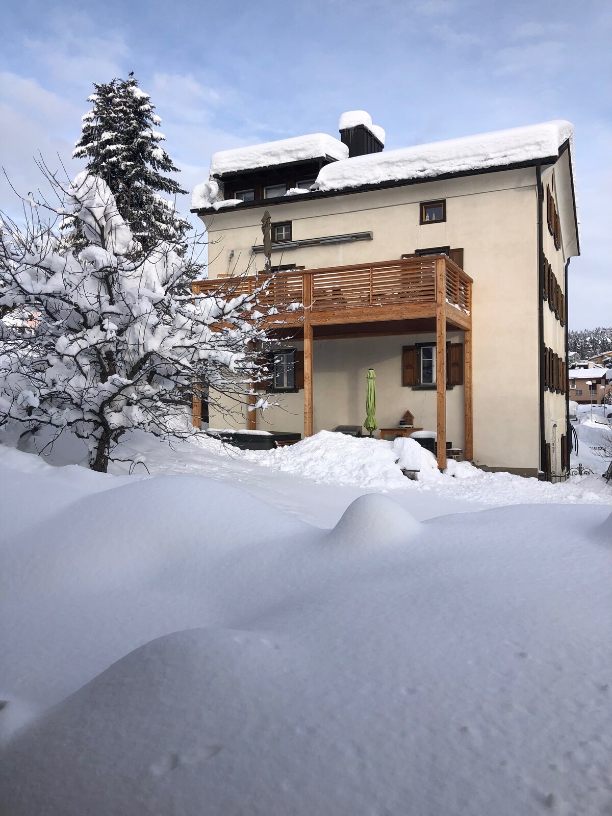 A renovated house is showcased under a blanket of snow, featuring a wooden balcony. Surrounding the home, snow-covered terrain and trees are visible, highlighting the serene winter landscape.