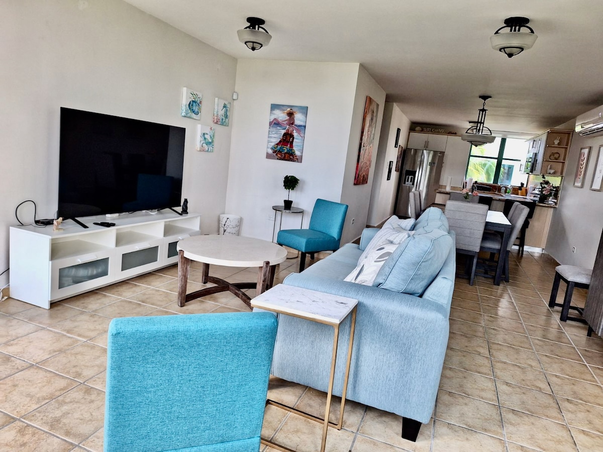 An open living area features modern, light-colored furnishings, including a spacious gray sofa and two teal chairs. A large flat-screen TV is mounted on the wall, and a round coffee table is positioned centrally. Natural light floods in through windows, creating a bright atmosphere.