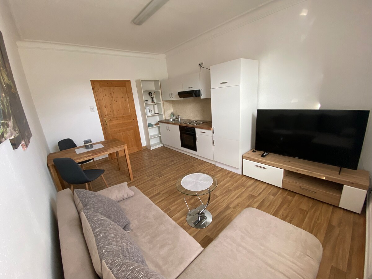 A spacious living area features a comfortable couch and a coffee table, with a TV mounted on a light wood unit. The adjoining kitchen includes modern appliances and a small dining table with two chairs, all set against a bright, neutral backdrop.