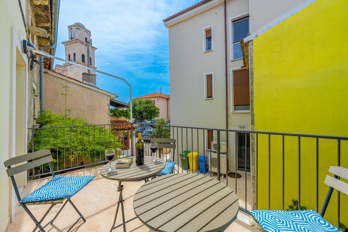 A balcony presents a casual seating area with a round table and two striped chairs, overlooking a picturesque street. Lush greenery is visible in the background, and the bell tower of St. Lovre's Church can be seen rising against a clear blue sky.