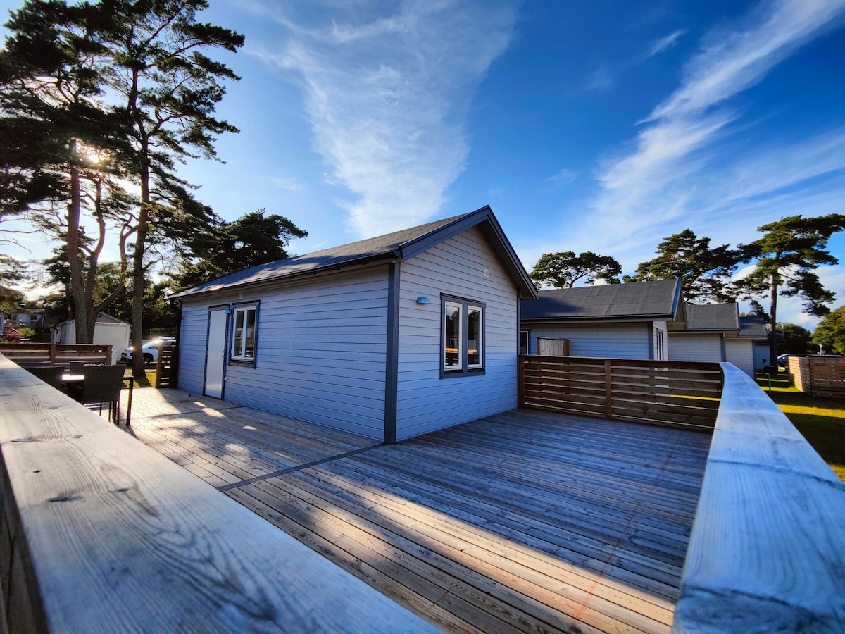 A light-colored cabin with large windows is set against a clear blue sky. A wooden deck surrounds the cabin, providing ample outdoor space. Trees are visible in the background, contributing to the natural ambiance of the area.