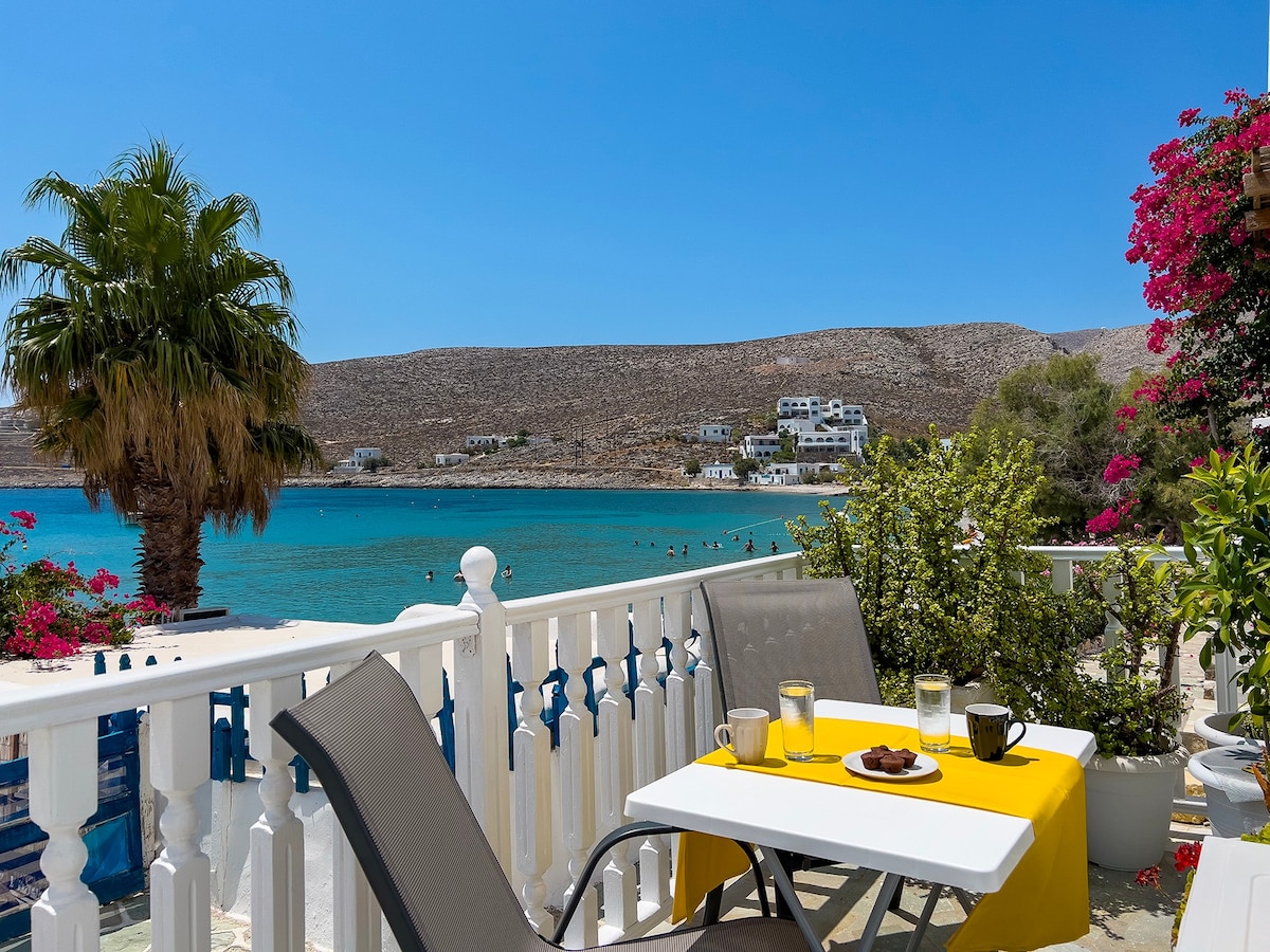 A charming outdoor terrace is framed by a white railing and lush greenery, offering a view of the turquoise sea. A table with a yellow tablecloth holds beverages and snacks, while comfortable chairs invite relaxation. Vibrant pink flowers add a pop of color to the scene.