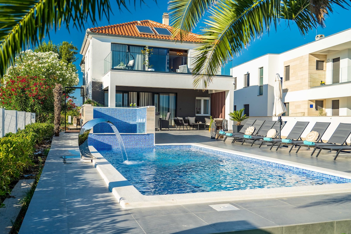 An outdoor space features a hydromassage pool surrounded by sun loungers and palm trees. The villa's white exterior is visible in the background, highlighted by a terracotta roof. Lush Mediterranean plants add vibrant greenery alongside the pool area.