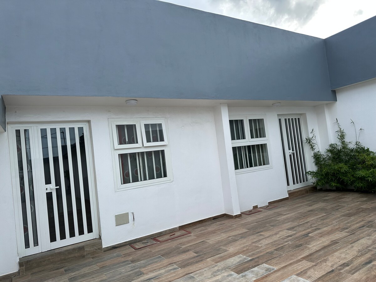 An exterior view showcases the modern design of the building, featuring three white-framed windows, two with prominent vertical bars. The light-colored walls contrast with the dark, slanted roof, while the tiled patio area offers a clean, spacious appearance, bordered by lush greenery.