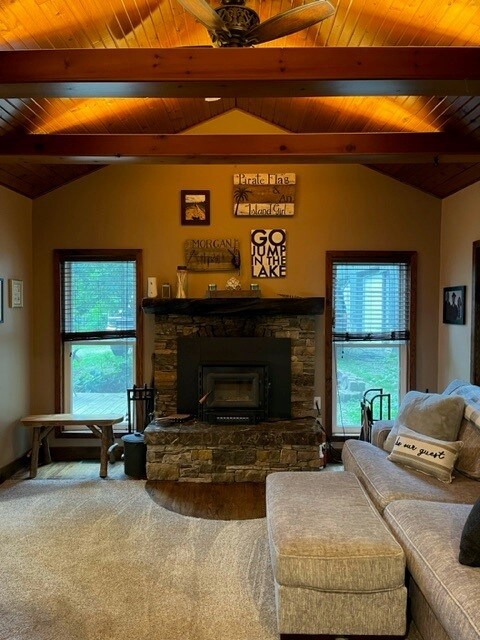 A cozy living area features a stone fireplace as its focal point, surrounded by wooden accents and large windows allowing natural light to fill the space. A textured gray sectional sofa faces the fireplace, while a small wooden table is positioned nearby.