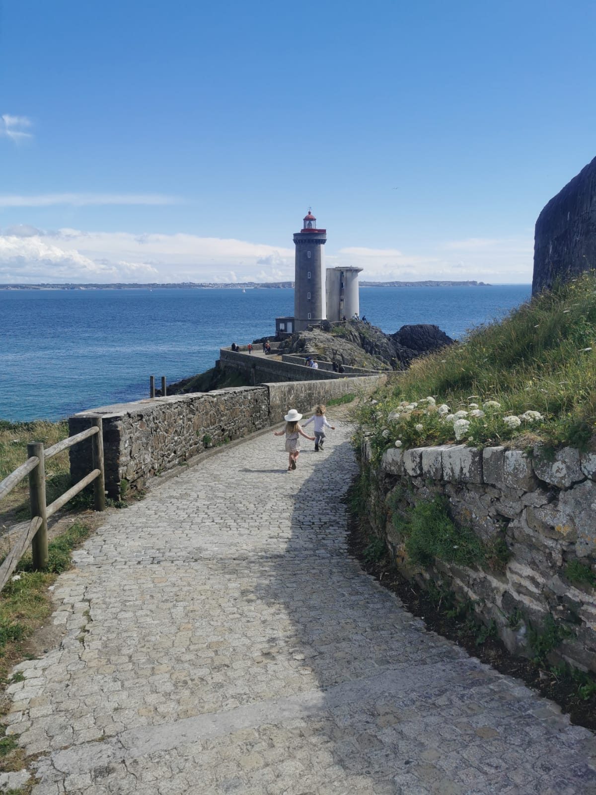 A scenic pathway leads towards a lighthouse overlooking the sea. Two children walk hand-in-hand along the cobblestone path, bordered by a grassy area and flowering plants, with the ocean visible in the background under a clear blue sky.