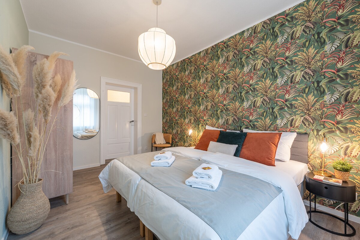 A bright and inviting bedroom features a stylish botanical wallpaper. Two beds are adorned with crisp linens and decorative pillows. A large round mirror reflects light, while a warm light fixture hangs from the ceiling. A clothing wardrobe and a cozy chair complement the space.