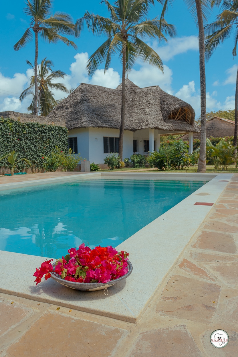 A refreshing swimming pool is surrounded by lush greenery and palm trees. In the foreground, a decorative bowl holds vibrant pink flowers, contrasting with the clear blue water. A thatched-roof structure is partially visible in the background, complementing the tropical setting.