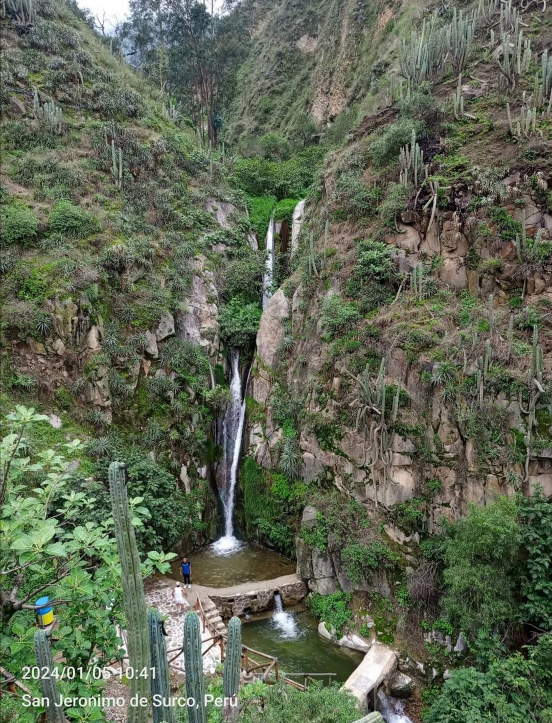 Two cascading waterfalls flow down rocky cliffs, surrounded by lush greenery and cacti. A stone pathway leads to the lower area, where visitors can be seen engaging with the natural surroundings. The scene conveys a tranquil and natural atmosphere.