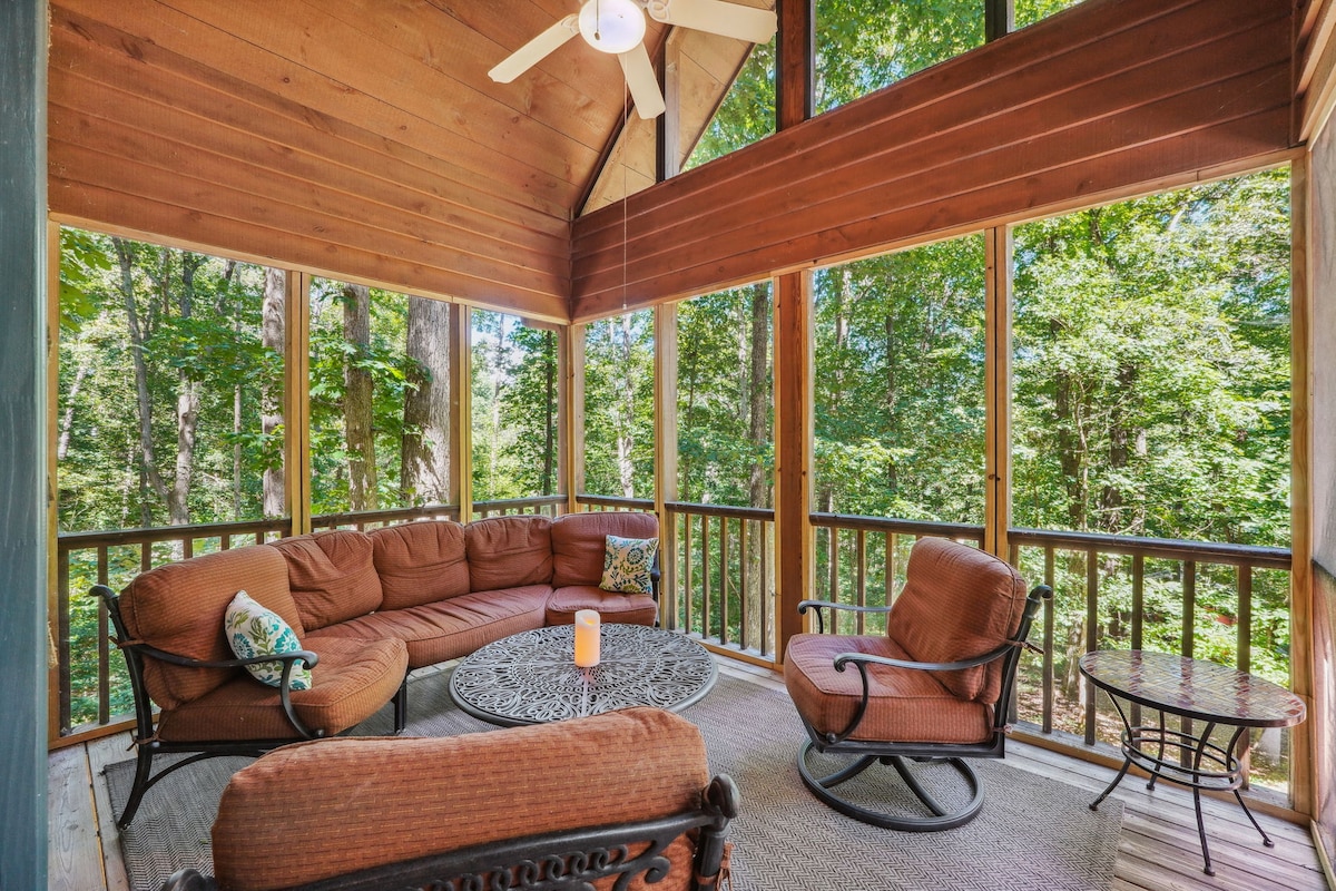 A screened porch features a circular brown sectional sofa surrounding a decorative table. Two coordinating lounge chairs provide additional seating. Natural light filters through the mesh screens, and a ceiling fan hangs above, enhancing the connection to the surrounding wooded landscape.