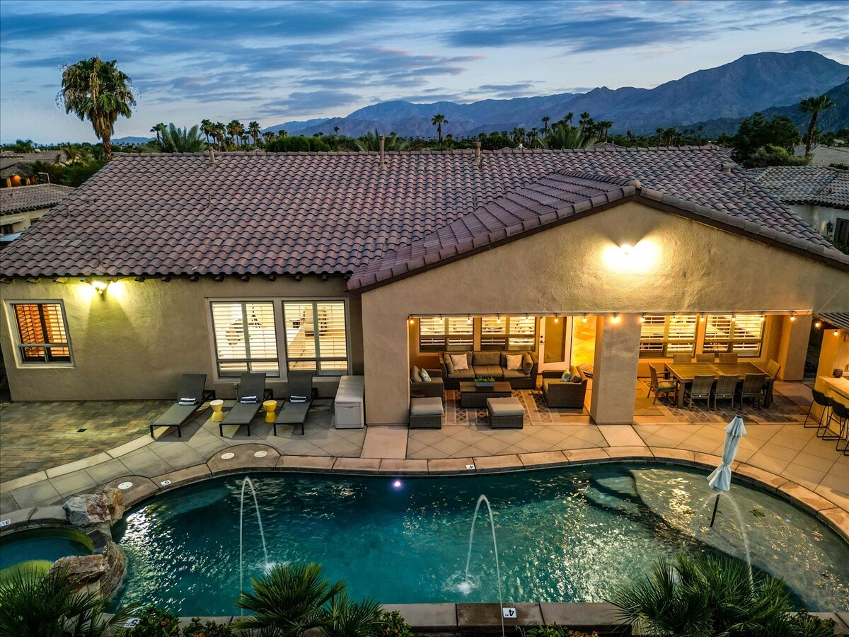 The villa's outdoor space features a turquoise pool with water fountains and comfortable lounge chairs positioned nearby. A covered seating area with multiple sofas and tables is visible through large glass doors. Mountain views and palm trees are framed in the background under a soft evening sky.