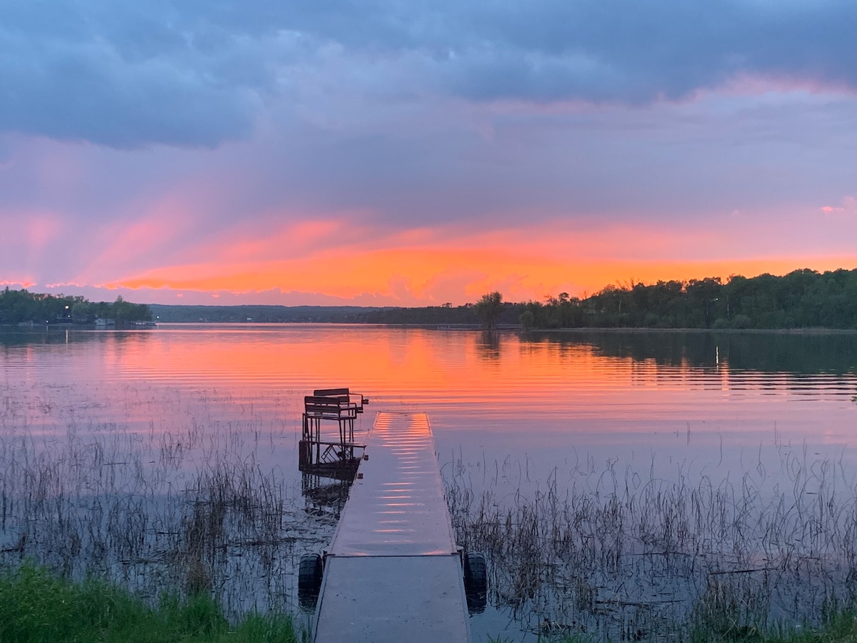 A serene lake view is captured at sunset, with vibrant colors reflecting off the calm water. A wooden dock extends into the lake, providing a focal point against the backdrop of softly lit clouds and distant trees.