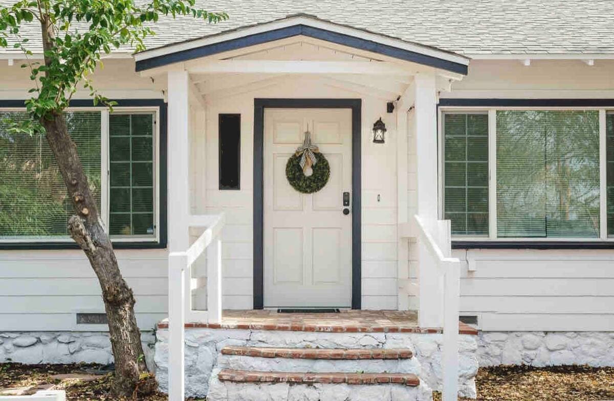 The entrance to the cottage features a charming door adorned with a wreath. Flanked by large windows, it is accessible via a set of brick steps leading to a welcoming porch with white railings.
