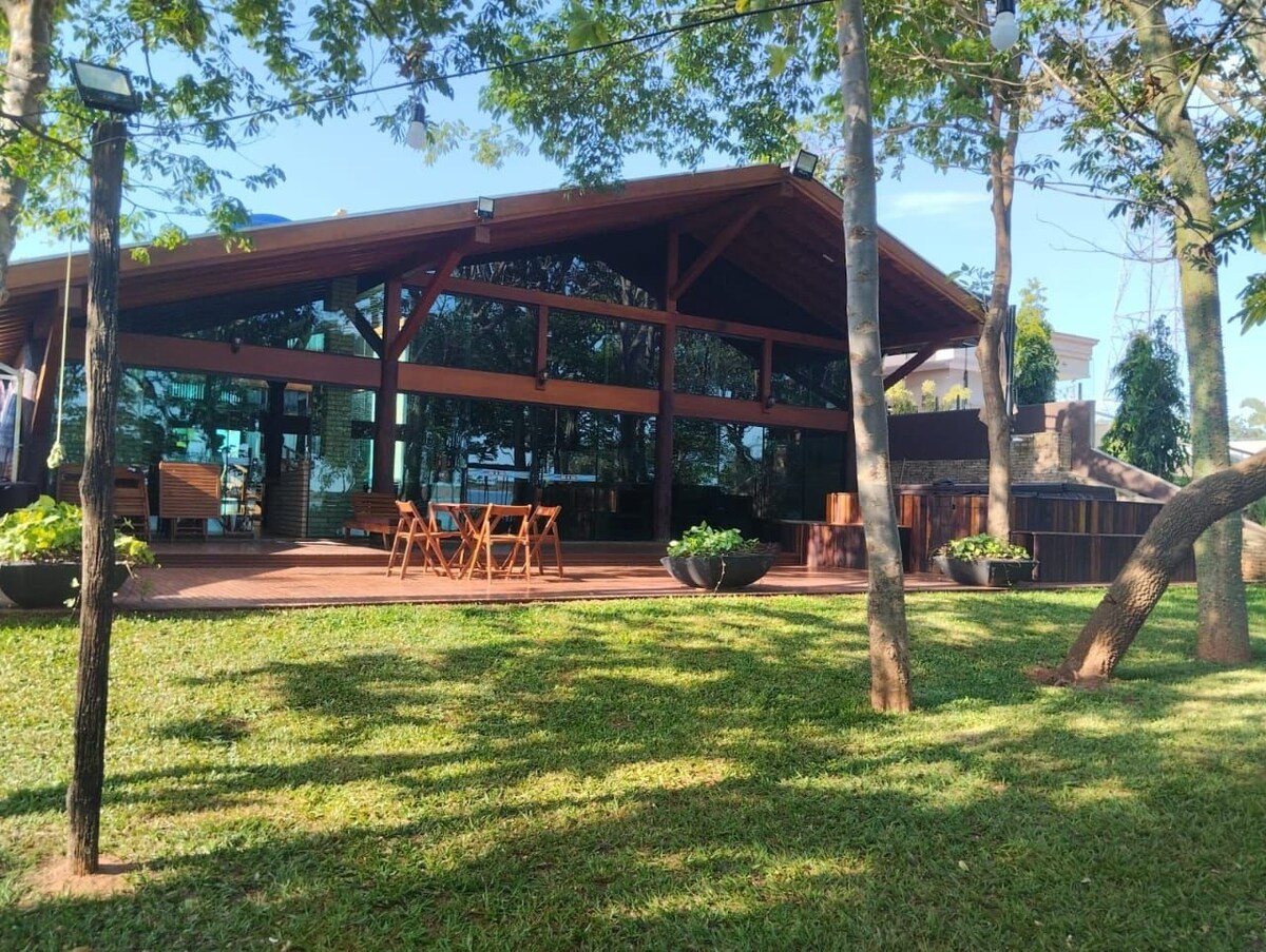 A spacious outdoor area features a wooden deck surrounded by lush grass and trees. A large structure with expansive glass walls provides ample natural light, with a seating arrangement of wooden tables and chairs visible. Notable landscaping includes decorative plant containers.