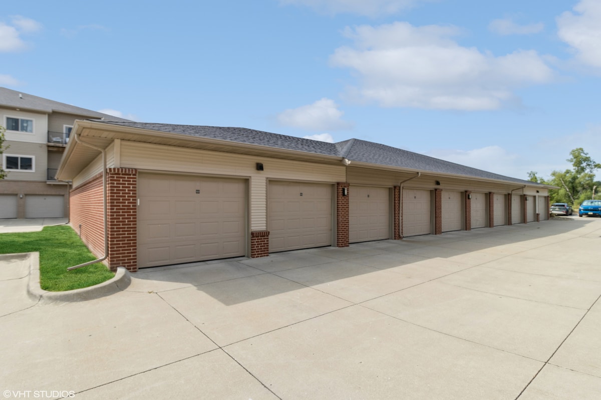 A row of private garages is displayed, featuring modern beige and brick exteriors. Each garage door is uniformly designed, creating a cohesive appearance. The surrounding paved area is well-maintained, with green grass lining the edges, contributing to a clean and organized environment.