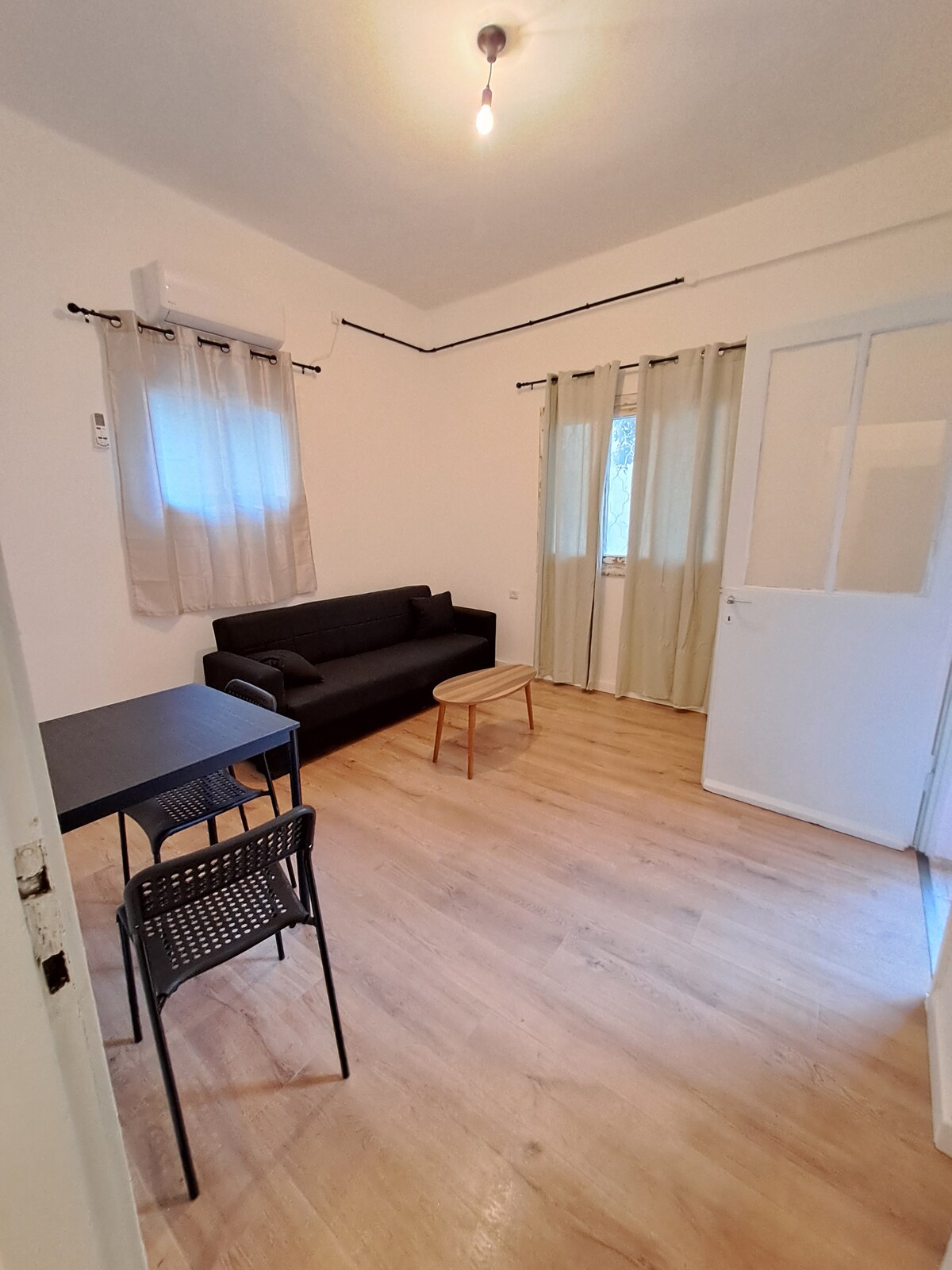A bright living area features a dark sofa positioned against a softly lit wall. A small wooden table and a chair are arranged nearby. Natural light filters through sheer curtains hanging over a window, enhancing the spacious feel of the room.