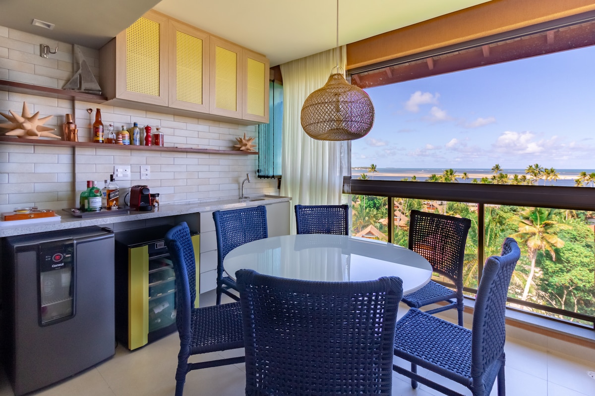 A well-equipped kitchen and dining area feature a round table surrounded by woven chairs. Modern appliances and shelving display kitchen items and decorations. Large windows provide a view of lush palm trees and the ocean beyond, enhancing the airy feel of the space.