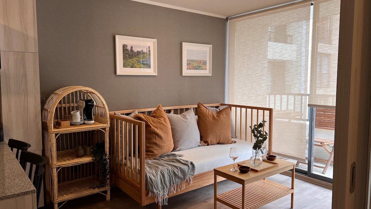A cozy living area features a light-toned sofa adorned with soft pillows and a textured throw. A natural wood coffee table sits in front, accompanied by a rattan shelf holding a coffee maker. Large windows allow ample natural light, with sheer shades partially drawn.