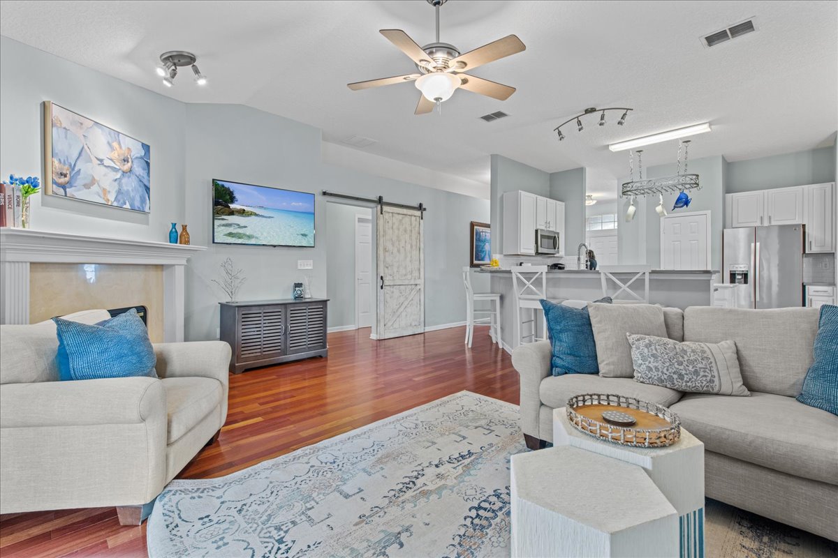 A bright open-concept living area features a light-colored sofa and armchair, accented with blue cushions. A modern ceiling fan is visible above, while a flat-screen TV is mounted on the wall. The polished wood floor complements the light walls and stylish decor.
