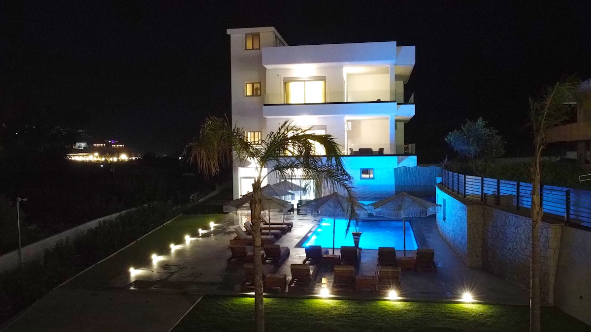 The modern three-story apartment building is illuminated at night, revealing expansive balconies. A swimming pool is surrounded by lounge chairs, with umbrellas providing shade. Soft garden lighting highlights the pathway and landscaping, enhancing the tranquil outdoor space.
