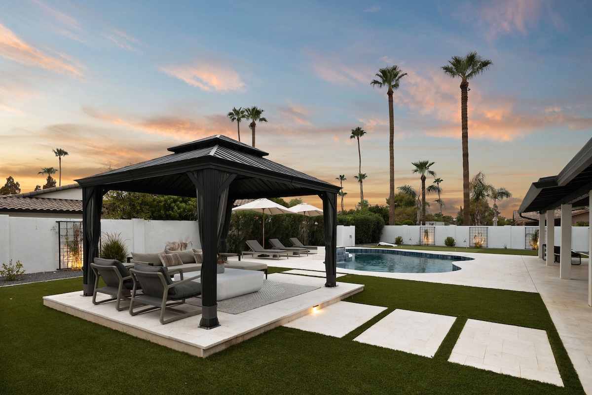 An outdoor lounge area is showcased under a black pergola, featuring comfortable seating and a sleek spa. The space is surrounded by palm trees, with a swimming pool and manicured grass providing a serene backdrop against a vibrant sunset.
