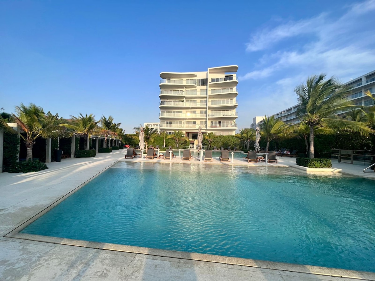A large pool is situated in the foreground of the image, surrounded by lush palm trees and lounge chairs. The spacious building is visible in the background, reflecting modern architectural design under a clear blue sky.