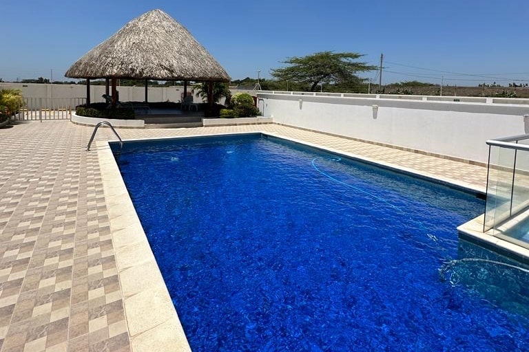 A spacious outdoor pool area is featured, with a vibrant blue pool surrounded by patterned tiles. A shaded gazebo with a thatched roof is visible near the pool, offering a space for relaxation. Lush greenery enhances the tranquil environment.