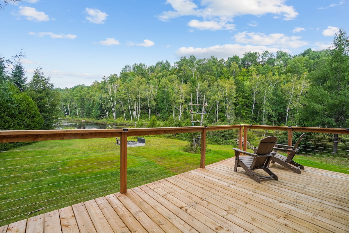 A spacious wooden deck features two outdoor chairs positioned to overlook a carpet of green grass, surrounded by trees. In the distance, a serene water view is visible, enhancing the natural setting of the outdoor space.