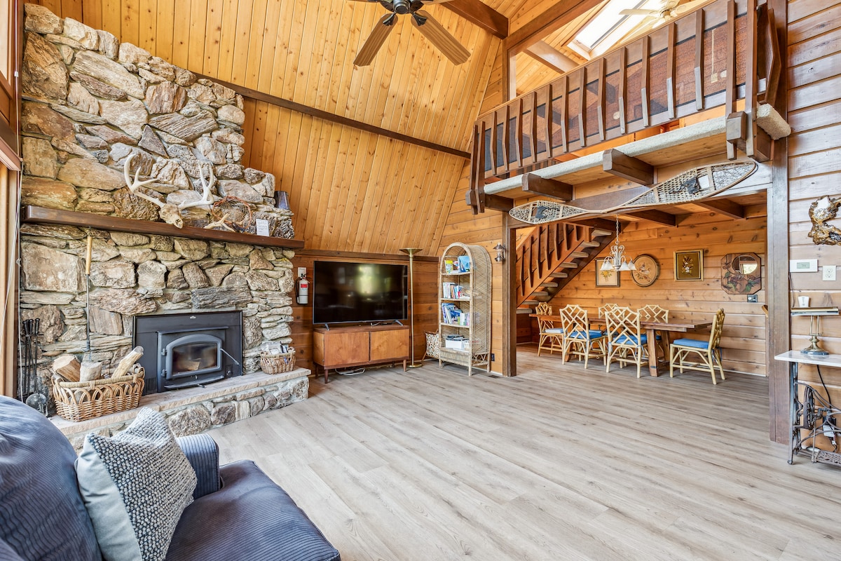 A spacious living area features a stone fireplace against a wooden wall, with a large television mounted above a wooden cabinet. A staircase leads to a loft area, while a dining table with multiple chairs is positioned nearby, allowing for gatherings.