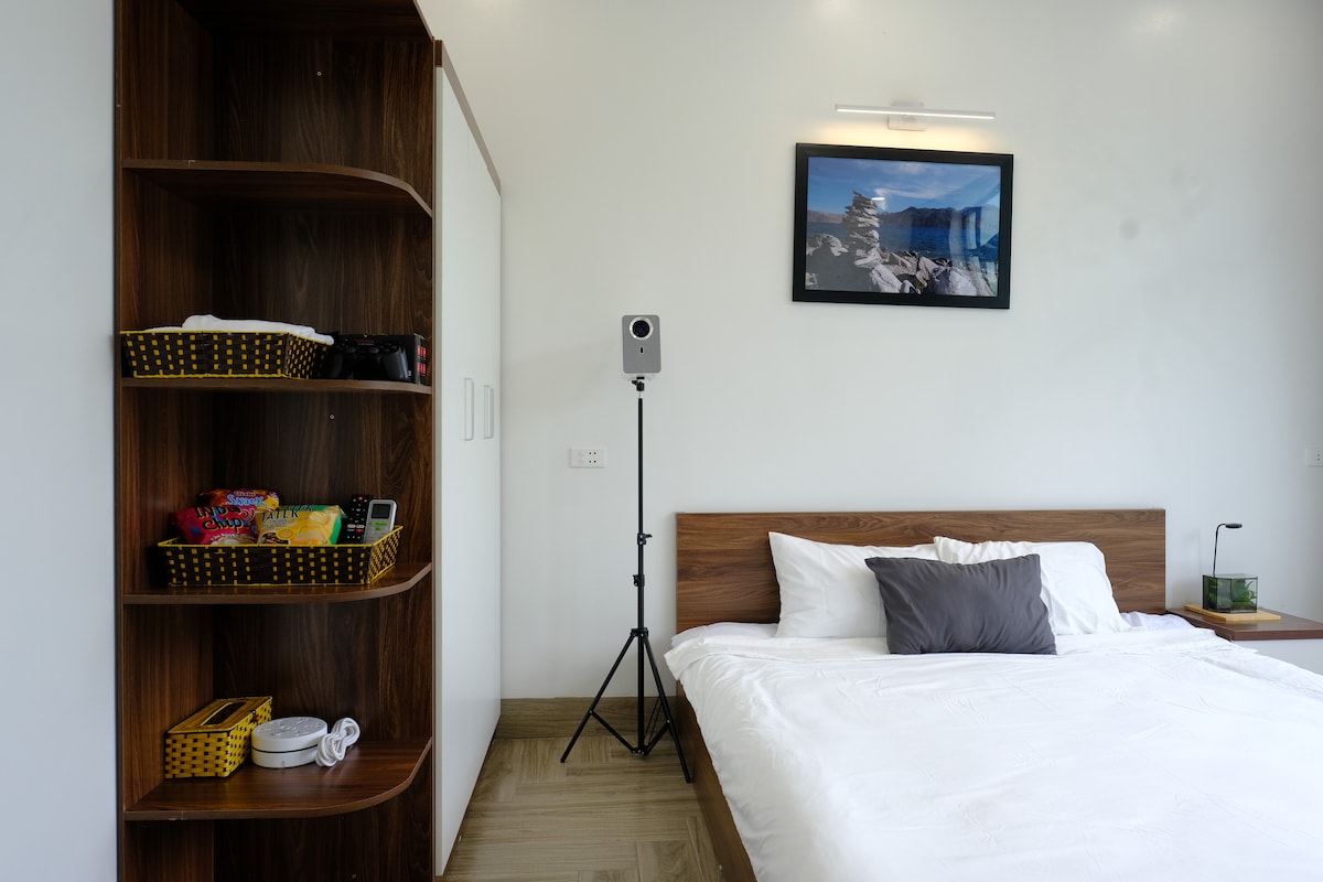 A bright and airy bedroom features a spacious bed with crisp white linens and a decorative pillow. A wooden shelving unit displays various items, including baskets filled with supplies. A framed photograph hangs above the bed, while a speaker stands beside it, adding to the modern decor.