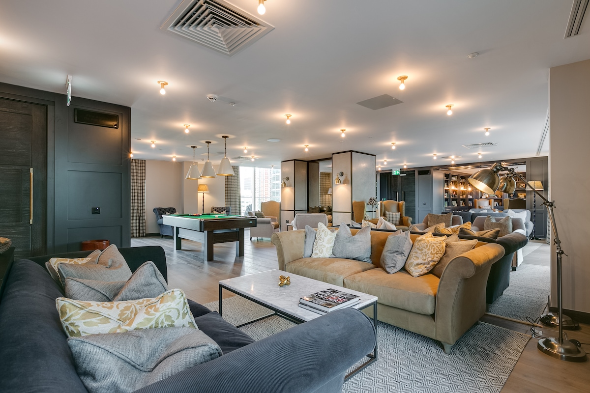A spacious communal lounge features a mix of comfortable seating arrangements, including sofas with decorative cushions. A billiards table is visible towards the back, complemented by ambient lighting from multiple fixtures throughout the area. A warm and welcoming atmosphere is created by the thoughtful layout.