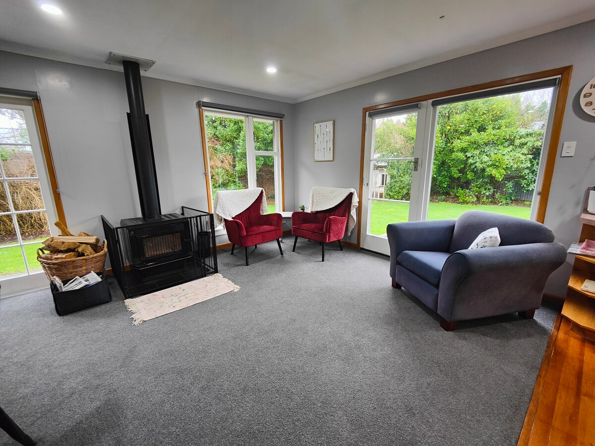 A cozy living area features two red armchairs and a blue sofa, arranged around a central wood-burning stove. Bright natural light fills the space through large windows overlooking a green backyard. The flooring consists of warm wooden elements, complemented by a soft gray carpet.