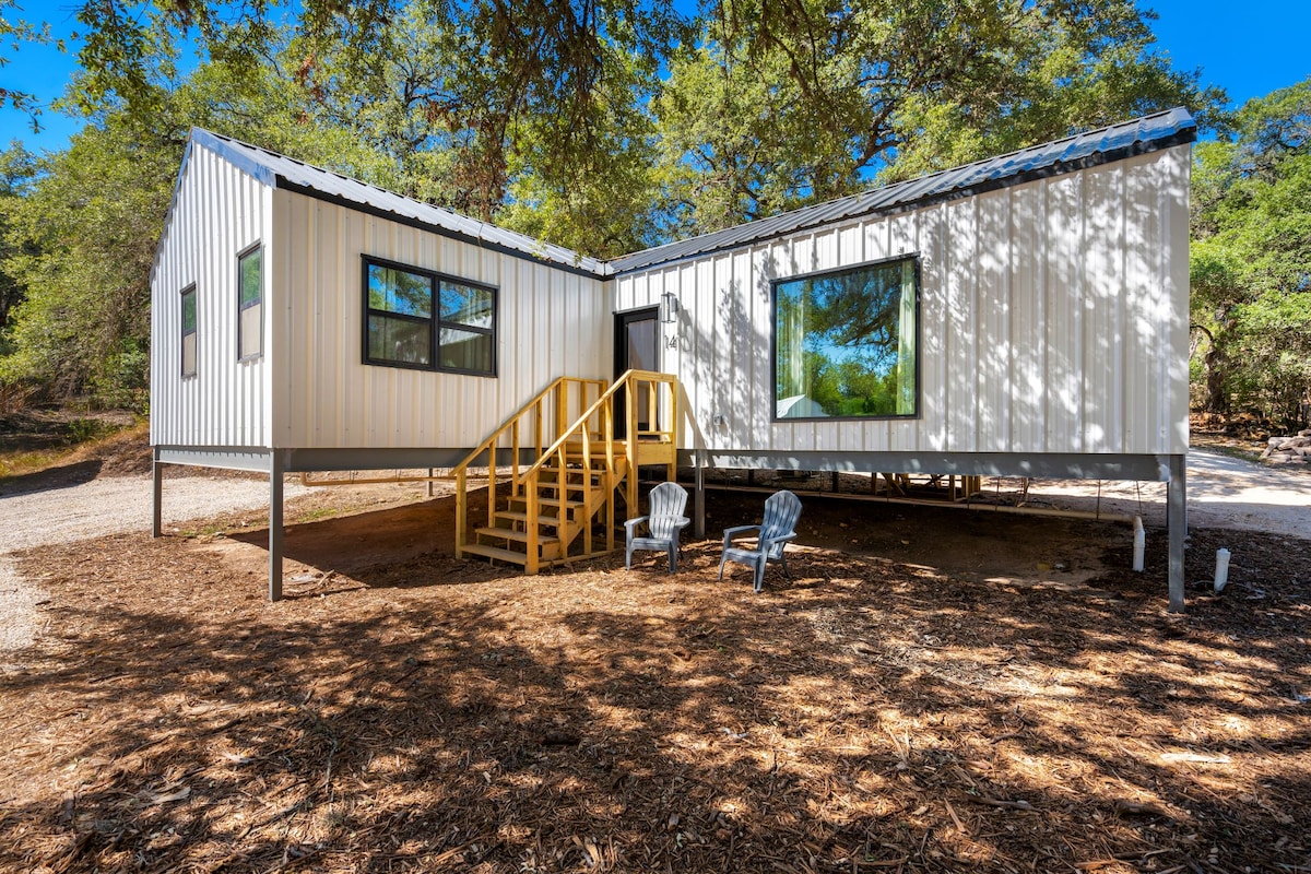 The modern cottage exterior features a combination of vertical metal siding and large windows that invite natural light. A yellow staircase leads to the entrance, and two Adirondack chairs are positioned on the ground, surrounded by natural landscaping.