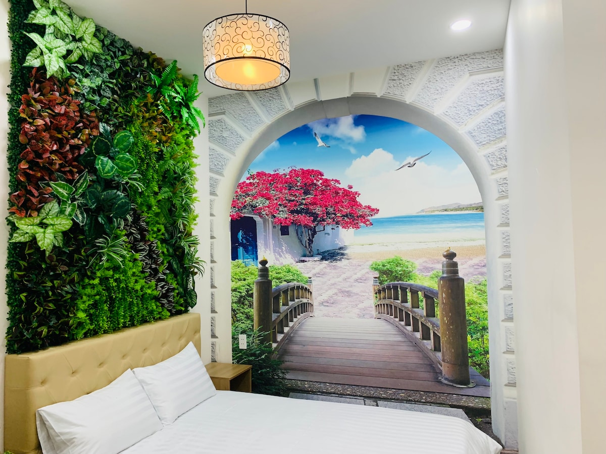 A serene sleeping area features a spacious bed with crisp white linens. A vibrant mural of a waterfront scene with lush greenery and red flowering trees serves as a stunning backdrop. A decorative light fixture hangs from the ceiling, adding a modern touch to the room.