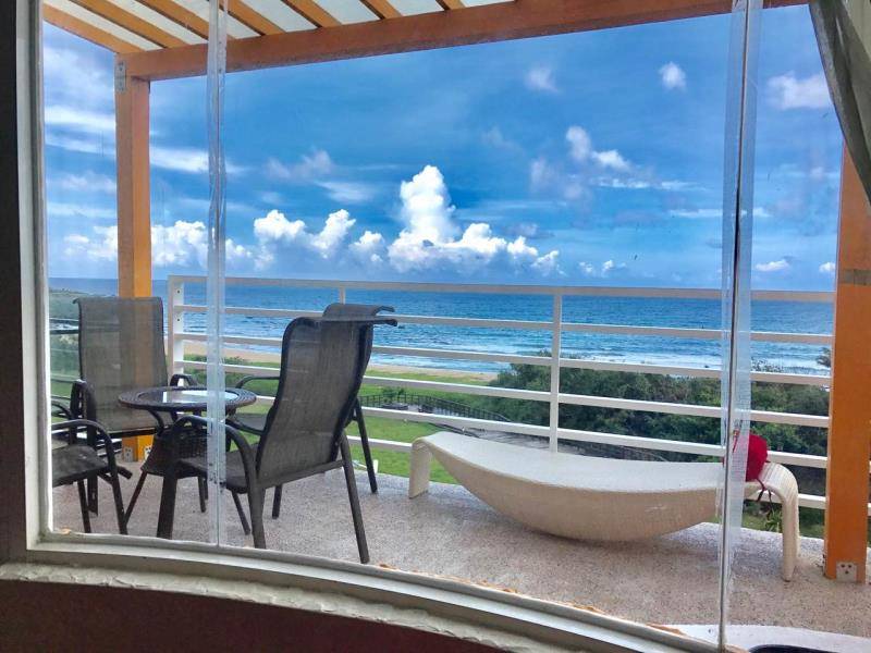 A view from the window captures a spacious balcony with outdoor furniture, including two chairs and a round table. The scene is complemented by a wide stretch of beach and ocean under a blue sky, creating a connection between indoor and outdoor spaces.