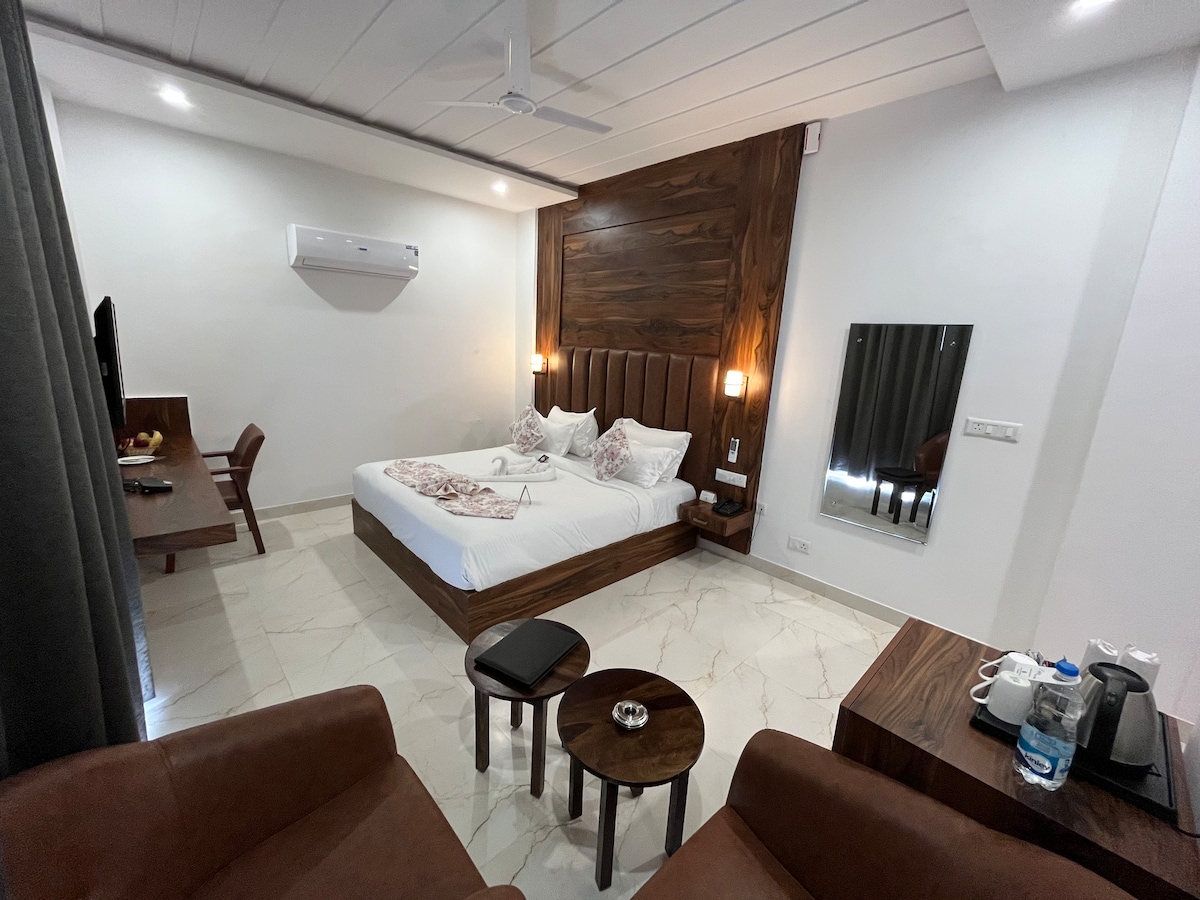 A sleeping area is designed with sleek wood paneling and soft lighting. A spacious bed is flanked by two bedside tables. A small seating area includes two brown chairs and a table, with a desk visible in the background. Amenities and a mirror are also present.