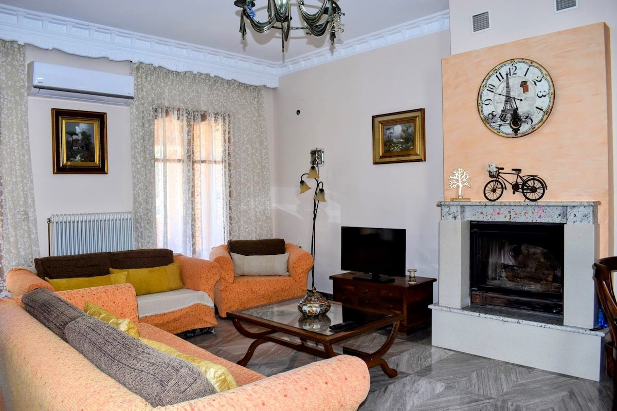 A living room showcases a cozy atmosphere with a mix of furniture, including a pair of sofas in warm tones and a coffee table. A decorative clock is mounted above the fireplace, while light filters through sheer curtains on a sunny day.