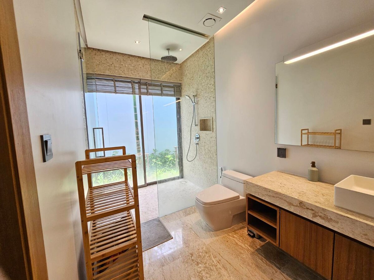 A modern bathroom features a frameless glass shower with a rainfall showerhead. Natural light flows in through large sliding doors, enhancing the stone wall texture. A wooden storage rack stands beside a sleek toilet, while a vanity with a minimalist sink offers functionality.