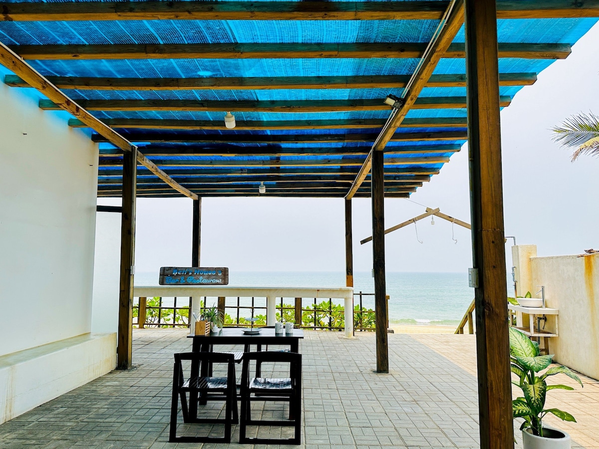 An open patio is featured under a blue canopy, offering views of the ocean. A simple wooden table is set with seat cushions, and the surrounding area is paved and well-maintained. The sound of the gentle waves can be imagined in the inviting space.
