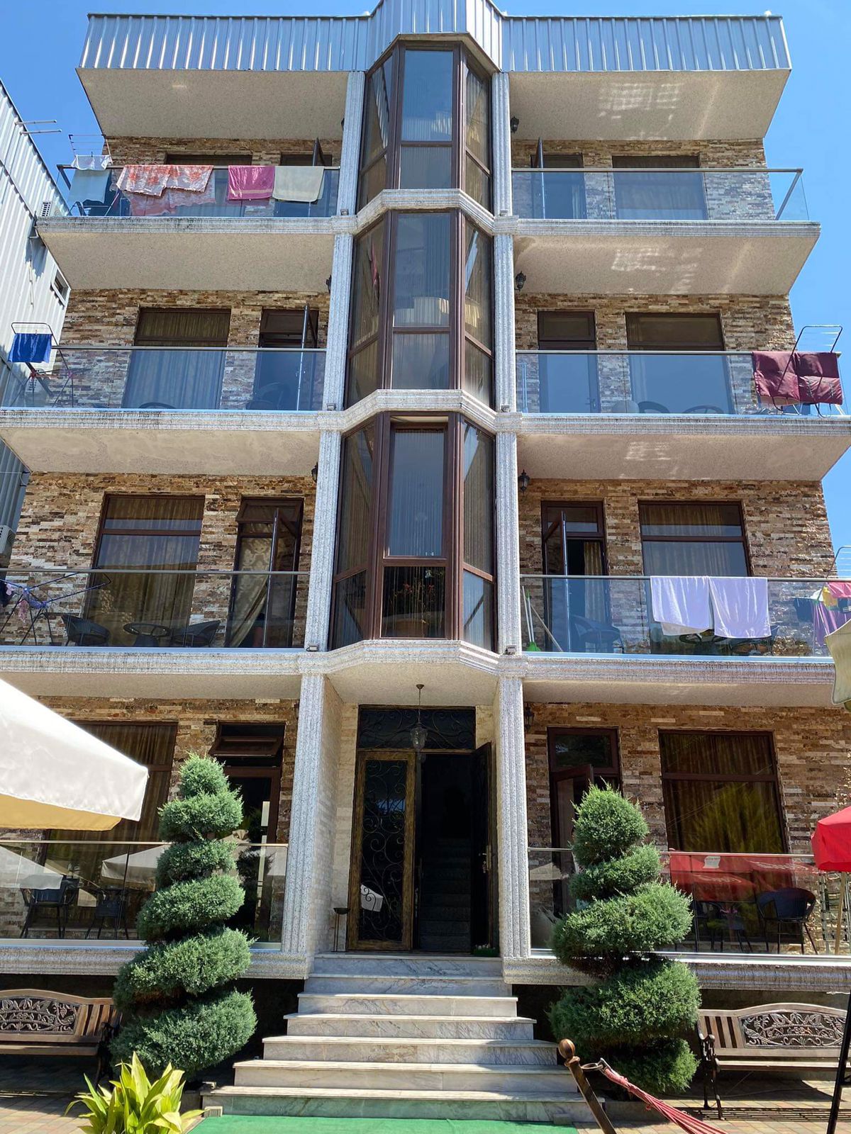 A three-story building features a modern design with large glass windows and a stone façade. Balconies are adorned with potted evergreen plants, and laundry is seen drying on the upper balcony. The entrance is accessed by a stone staircase, leading to a covered seating area.