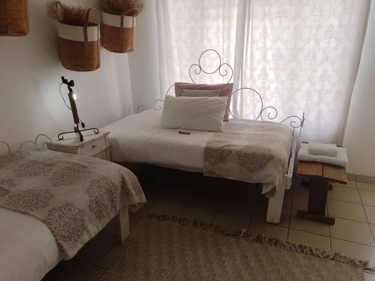 A well-lit sleeping area features two beds adorned with soft bedding and decorative throws. A small nightstand holds a lamp, while towels are neatly arranged on a nearby stool. Natural light filters through sheer curtains, creating a calm and inviting ambiance.