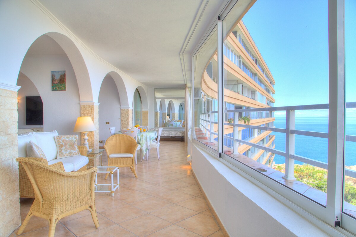 A spacious lounge area is featured with comfortable seating arrangements, including wicker chairs and a white sofa. Large windows offer expansive views of the ocean, allowing natural light to illuminate the space. The tiled floor complements the bright atmosphere.